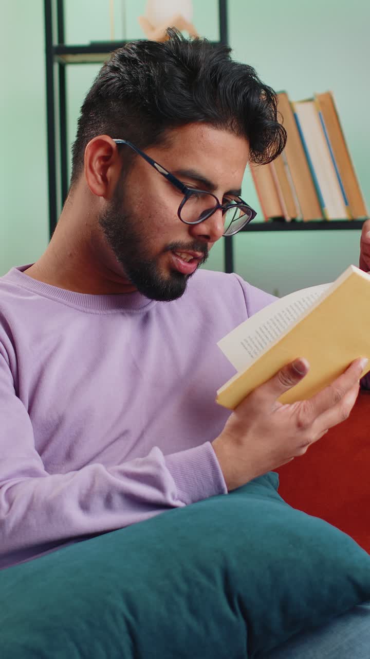 Indian man reading interesting book turning pages smiling enjoying literature taking a rest on sofa