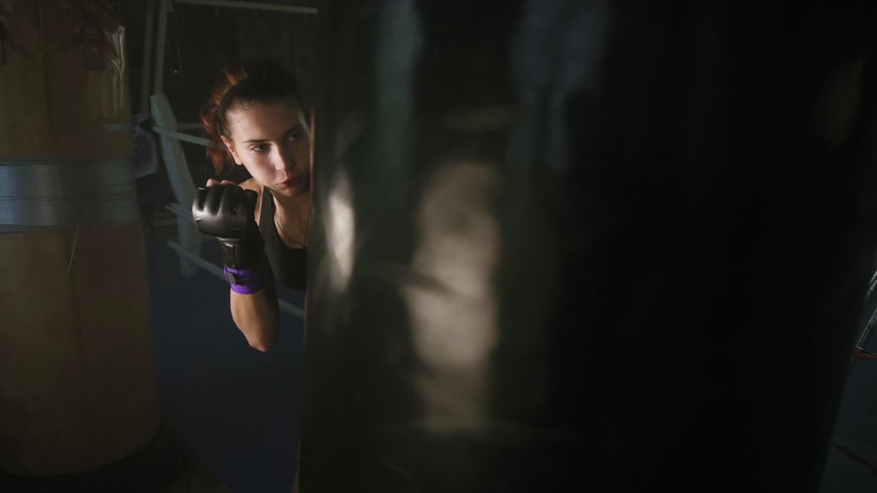 Athletic female boxer in gloves hitting boxing bag with her fist while training in a dark fitness studio with smoke. Slow Motion