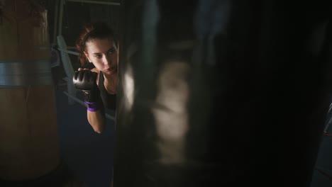 Athletische-Boxerin-In-Handschuhen-Schlägt-Mit-Der-Faust-Auf-Den-Boxsack,-Während-Sie-In-Einem-Dunklen-Fitnessstudio-Mit-Rauch-Trainiert.-Zeitlupe