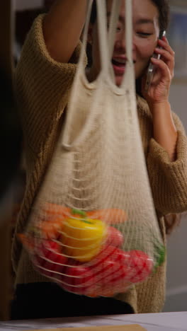 video vertical de una mujer hablando por teléfono móvil llegando a casa de las compras de alimentos desempaquetando una bolsa de verduras frescas en el mostrador en la cocina 1