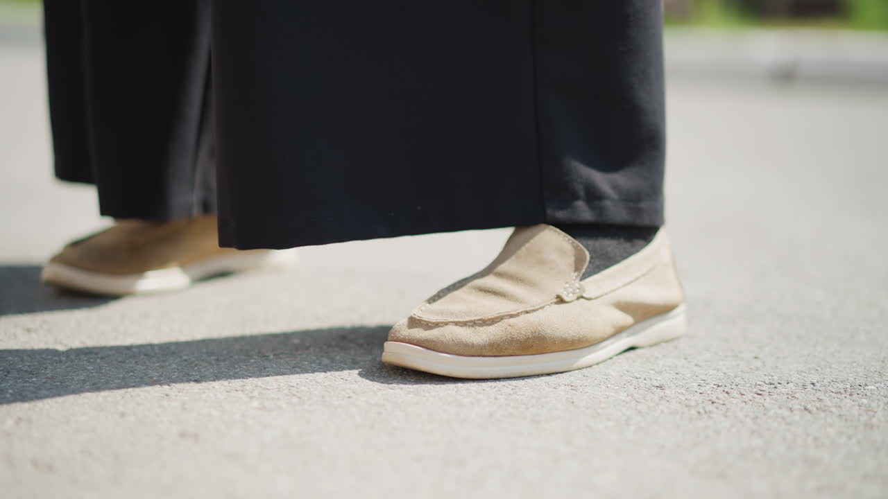 Close up of lady legs moving slightly on sunlit pavement, wearing black pants and brown shoes, subtle shifting motion reflects nervousness, curiosity, and emotional unease through soft light