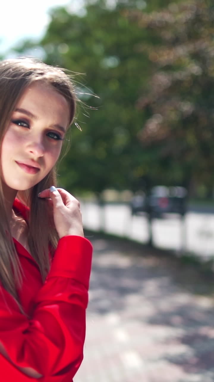 Lovely girl in red dress. Beautiful young model is standing outdoors and posing on camera. Portrait of a pretty woman on the green trees background. Vertical video