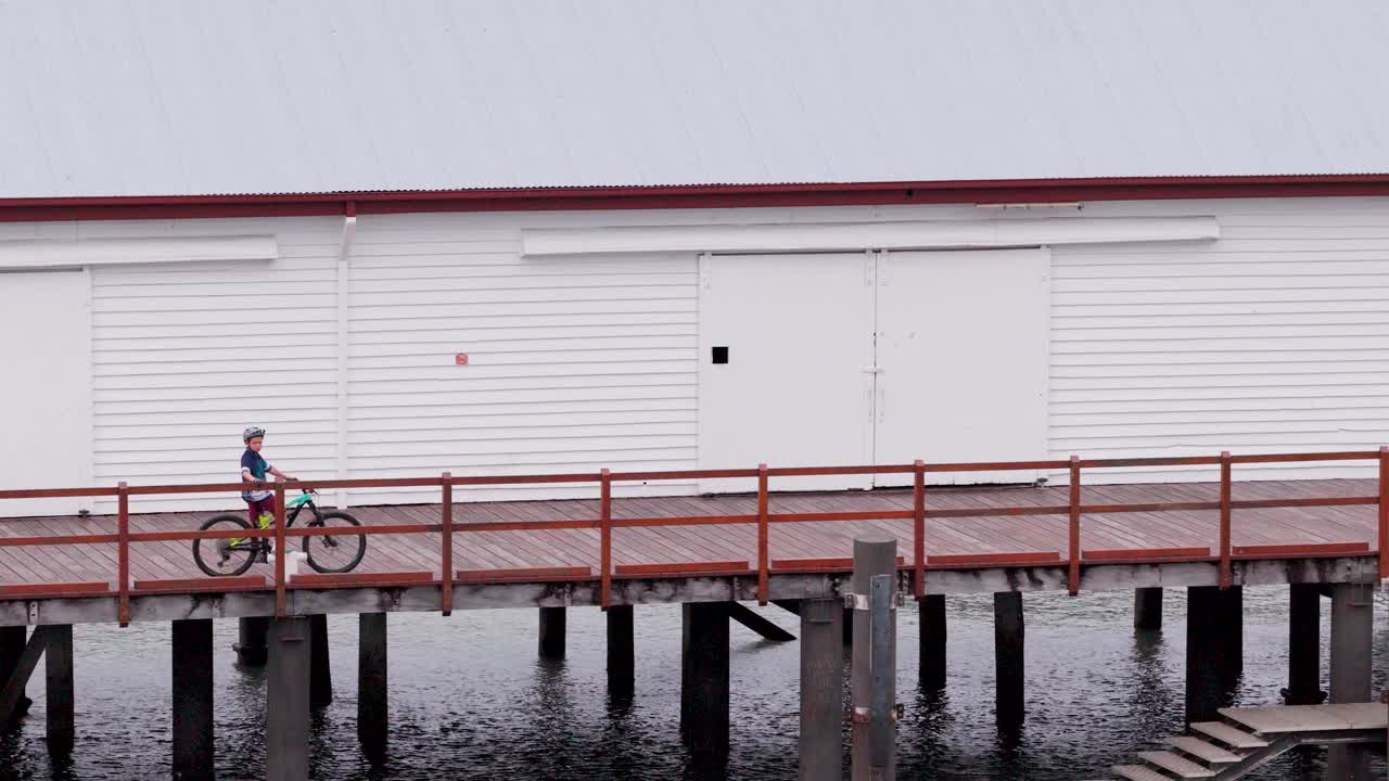 A child rides a bicycle along a wooden pier in Port Douglas, Australia. The scene captures a serene coastal environment with calm waters
