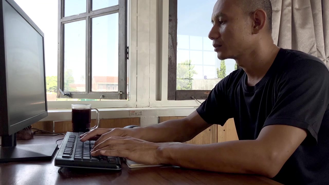 Nahaufnahme eines Mannes in schwarzem Hemd, der im Homeoffice arbeitet und sich konzentriert auf seiner Laptop-Tastatur an einem Holzschreibtisch tippt.