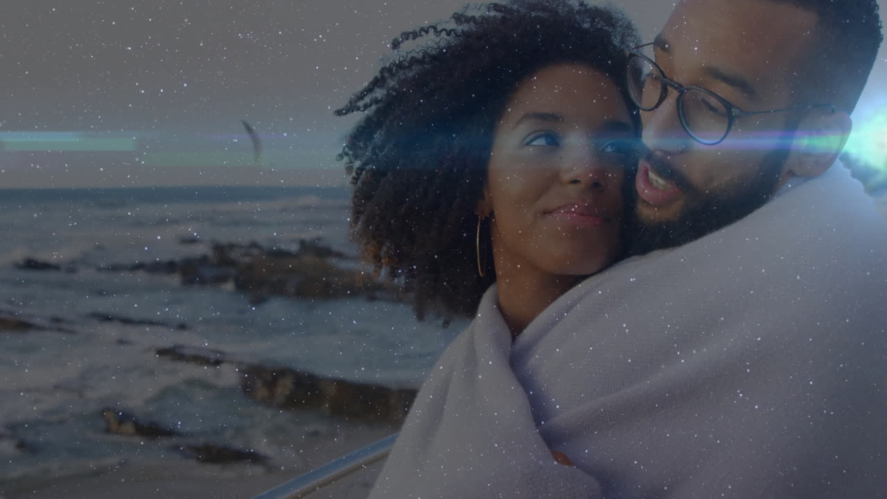 Coppia che si abbraccia sotto una coperta su una spiaggia rocciosa al crepuscolo per la salute, con icone a forma di cuore animate fluttuanti.