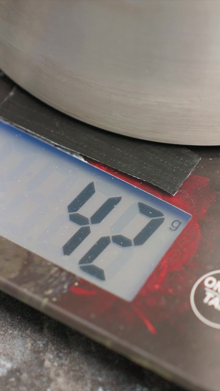 Primo piano di bilancia digitale che mostra il peso in grammi mentre un oggetto vi è posizionato sopra. Il display digitale mostra numeri che cambiano indicando la misurazione della massa, con materiale metallico e testurizzato visibile.