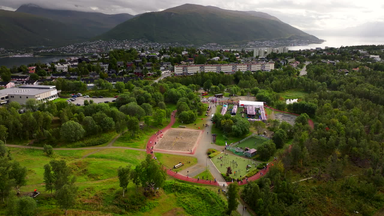 Relay for Life charity event in Tromsø, people walking on track, tents, and stage, with city and mountains in background, Norway. Aerial drone
