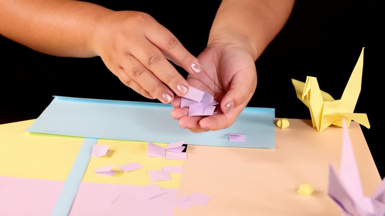 Hands gather small purple paper squares on pastel surface, surrounded by origami cranes, studio lighting