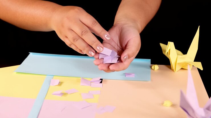 Manos recogiendo pequeños cuadrados de papel morado sobre una superficie en tonos pastel, rodeados de grullas de origami e iluminación de estudio