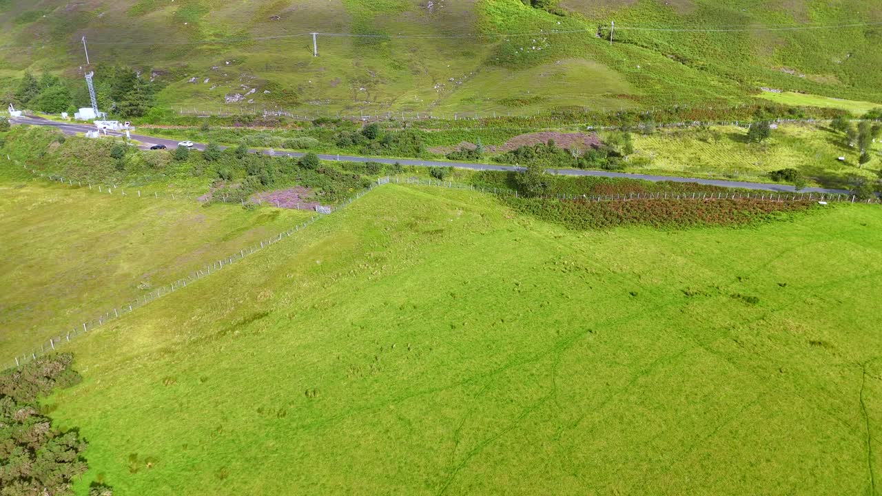 Drone footage glides above lush green fields, a winding rural road, and scattered farm structures in the Highlands, under soft natural daylight