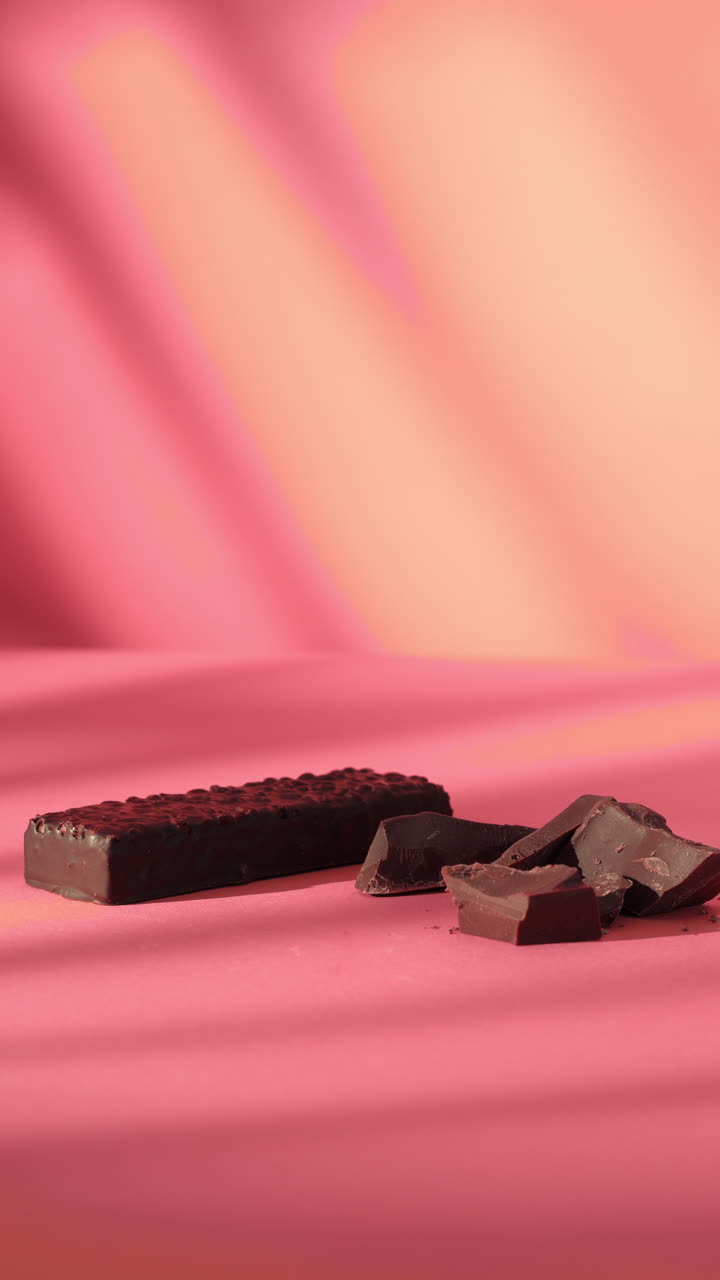 Close-up of a hand gently placing a dark chocolate protein bar next to broken chocolate pieces on a pink surface. Soft lighting and minimal background emphasize clean and modern food presentation.