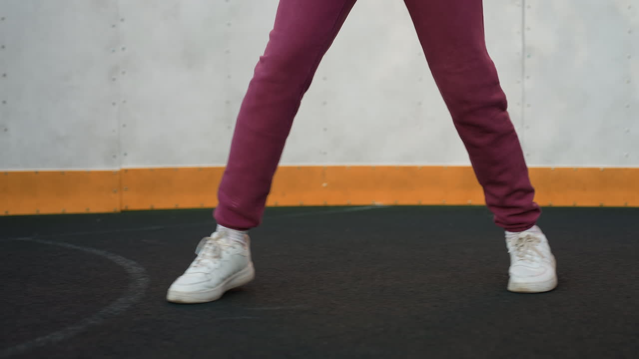 Leg view of urban woman jumping around round sports court wearing white sneakers and purple joggers attire under clear sky with concrete wall and fence background vibrant movement focus on feet