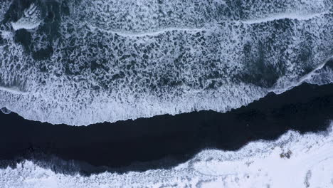 Toma-Aérea-De-Arriba-Hacia-Abajo-De-Las-Olas-Del-Océano-Que-Alcanzan-La-Arena-Negra-De-La-Playa-Volcánica-Cubierta-De-Nieve-Y-Hielo-En-Islandia