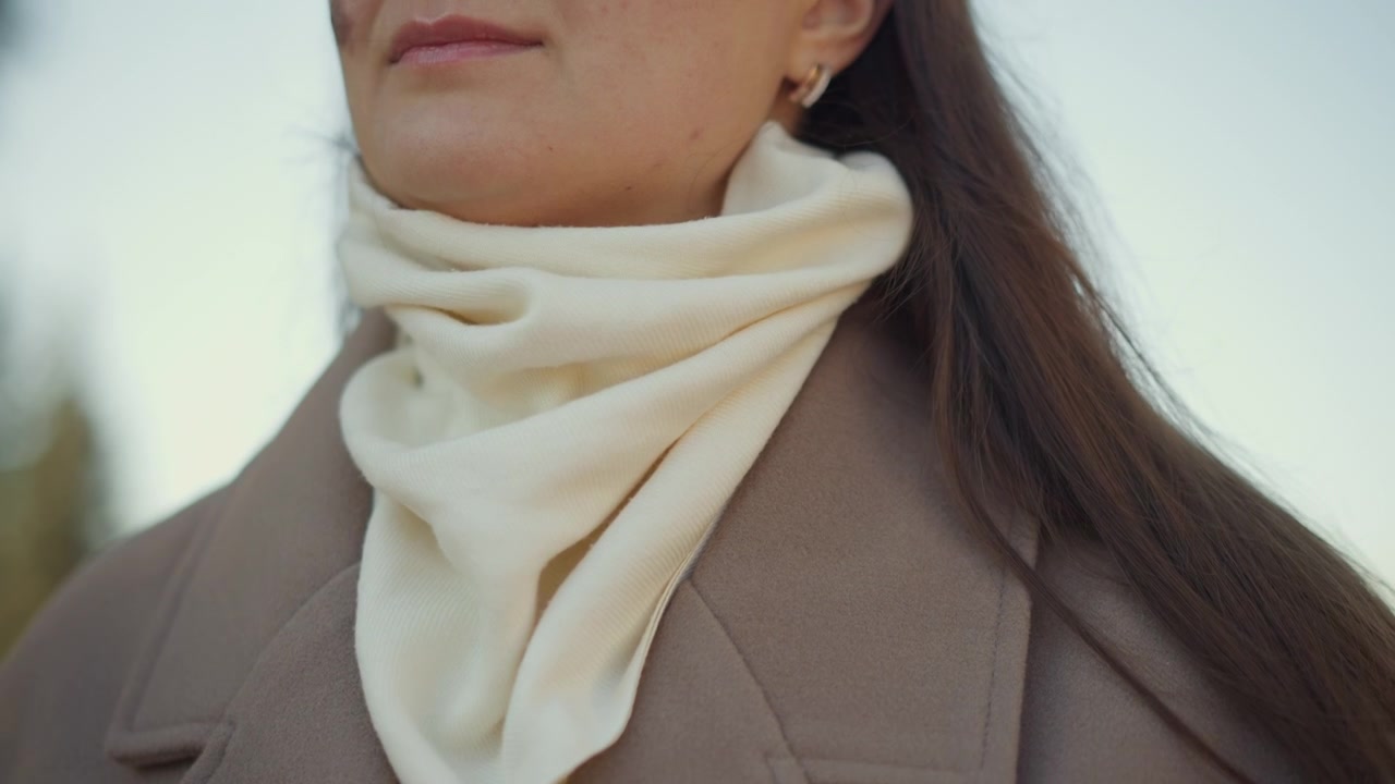 Primo piano di una donna che si sistema la sciarpa color crema intorno al collo mentre indossa un cappotto marrone, con luce soffusa e un'espressione calma. Il movimento della mano è visibile mentre la donna sistema il tessuto con lunghi capelli scuri.