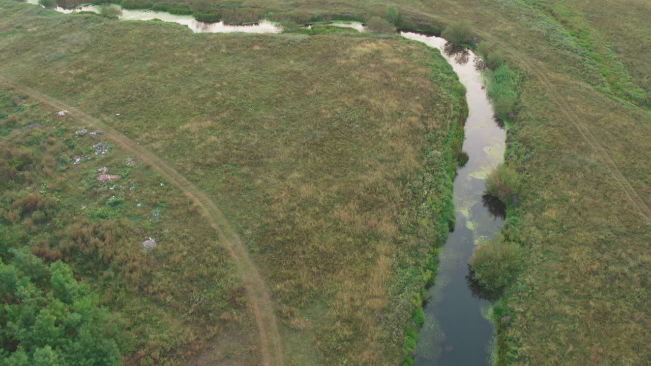 Aerial drone shot tracks slowly over a narrow, winding stream flowing between grassy, agricultural fields and the dark green edge of a forest, suggesting isolation and nature