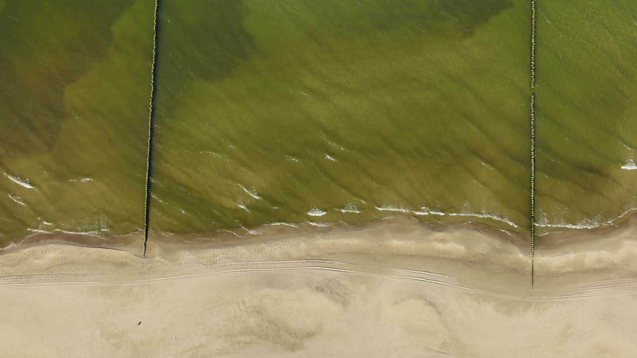 Top-down aerial view of a serene beach with clear green sea waters gently lapping against a sandy shoreline, marked by subtle wave patterns and a single trail across the sand