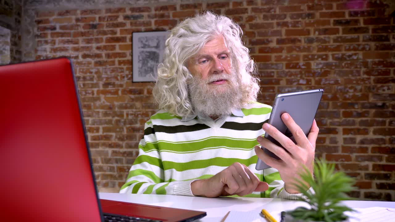 Happy old caucasian man with white magnificent beard and wavy hair is demonstrating blue screen on his tablet while sitting indoor, red background