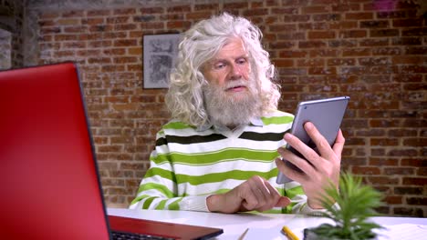 feliz anciano caucásico con barba blanca magnífica y cabello ondulado está demostrando pantalla azul en su tableta mientras está sentado en el interior, fondo rojo