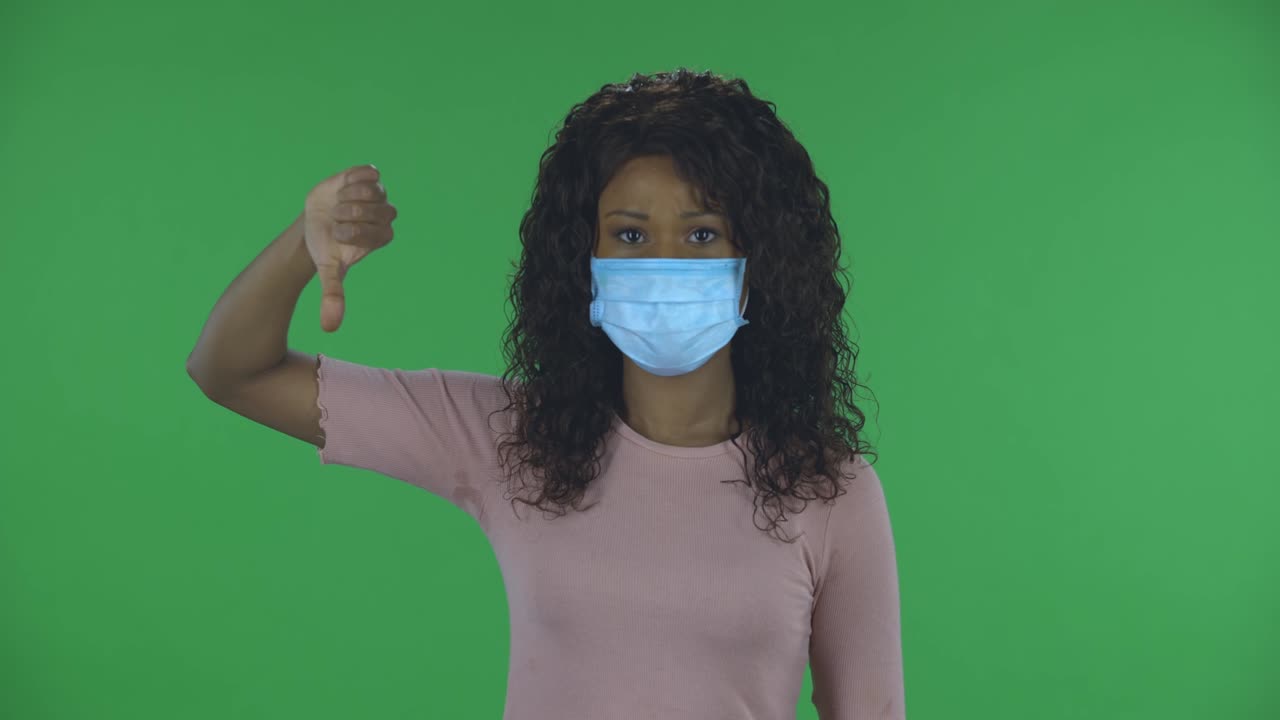 Portrait of beautiful african american young woman in medical protective face mask looking at camera and shows fingers down on both hands. Burning brunette with wavy hair in jeans and a beige blouse on a green screen in the studio. Health Protection