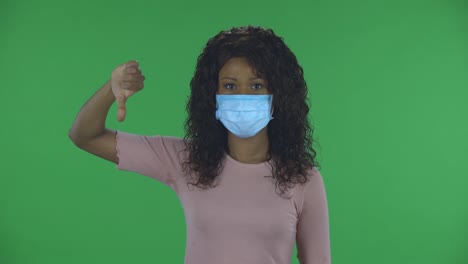 portrait of beautiful african american young woman in medical protective face mask looking at camera and shows fingers down on both hands. burning brunette with wavy hair in jeans and a beige blouse on a green screen in the studio. health protection