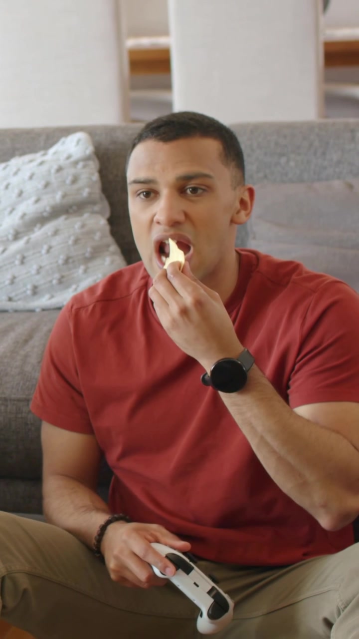 Vídeo vertical: Homem de camiseta vermelha virando-se para pegar batatas fritas na sala de estar, segurando um tablet após um alerta.