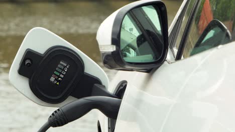 An-electric-car-charging-on-a-city-street,-a-cable-connected-to-a-car,-future-of-the-automobile,-clean-sustainable-energy,-overcast-day,-medium-shot,-water-on-the-background