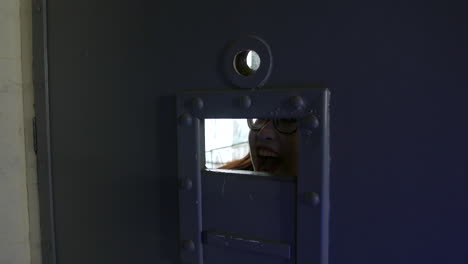 a woman is screaming while peeking through a small hatch in a prison or jail cell door