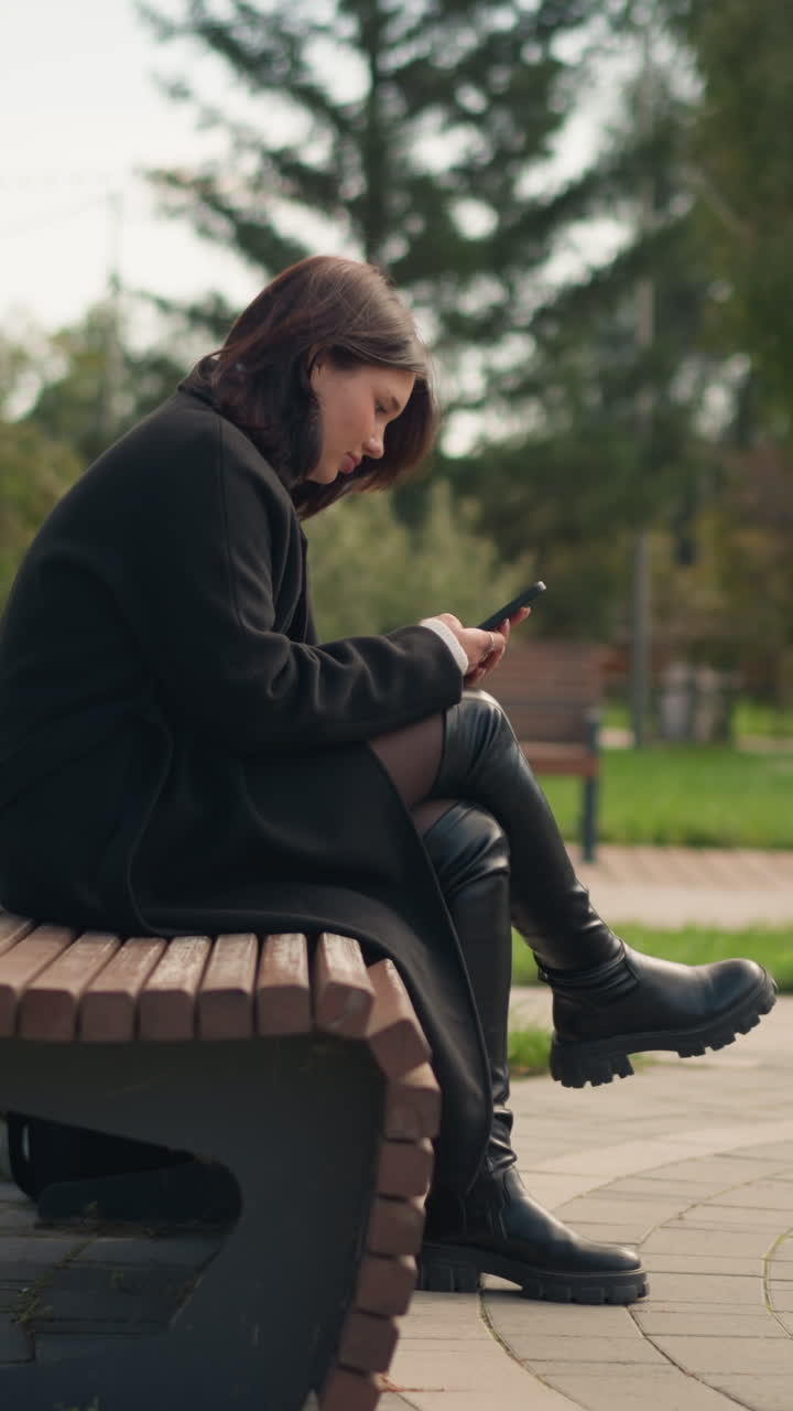 mujer sentada en un banco del parque usando un teléfono inteligente, vestida con abrigo y botas negras, rodeada de árboles verdes y exuberantes, atmósfera al aire libre relajada en un parque urbano, vista borrosa de una persona en una silla giratoria