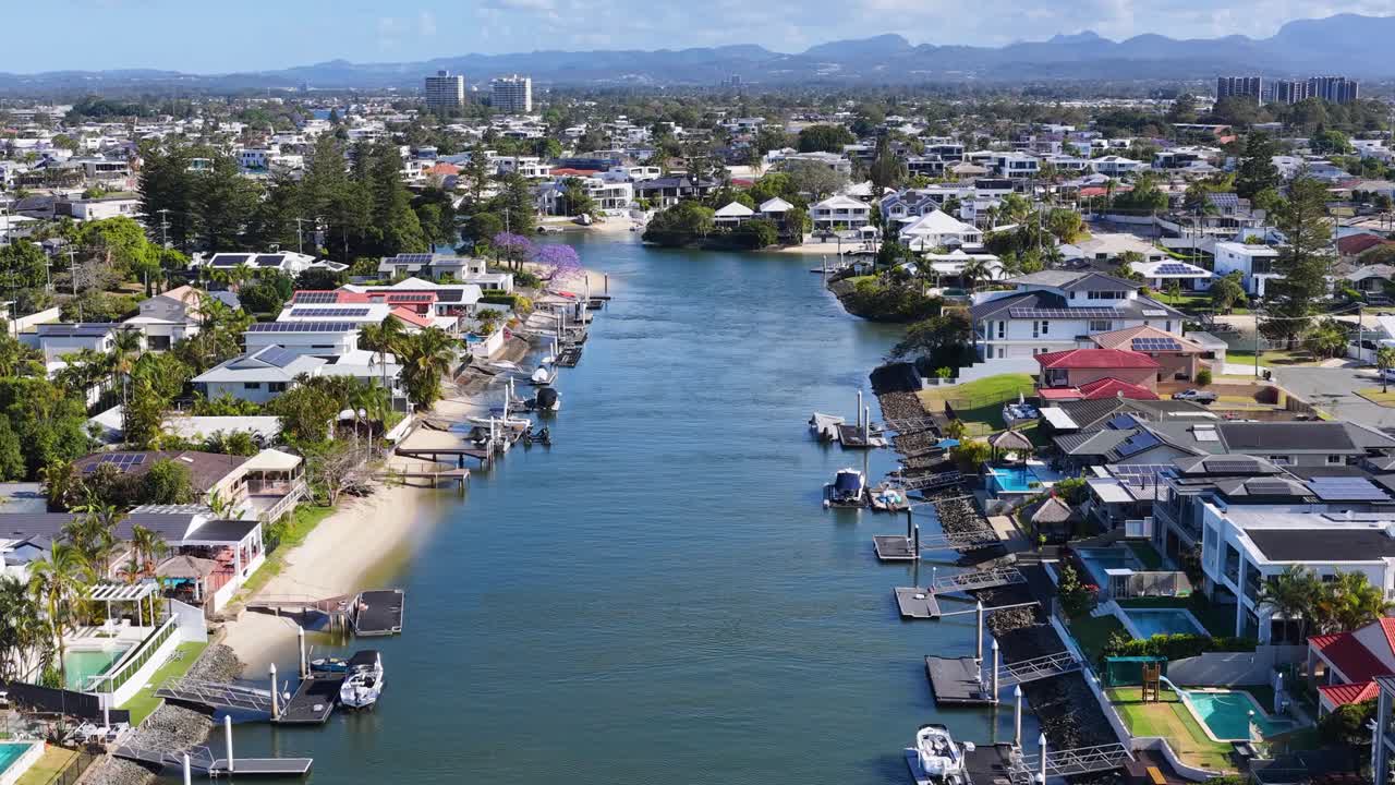Drone glides above canal, luxury waterfront houses, docks, and lush greenery in bright daylight