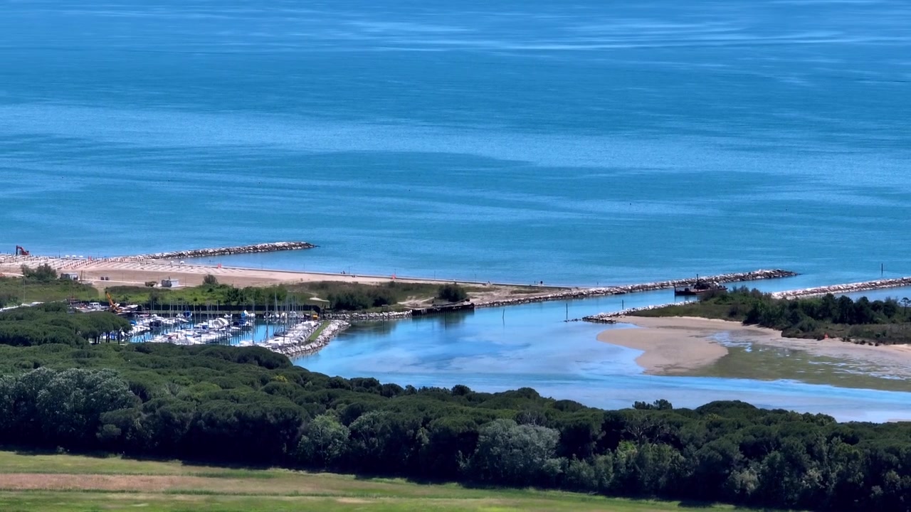 Ampia ripresa aerea con drone in movimento che rivela l'ingresso del canale del piccolo porto marittimo e i moli delle barche vicino alla foce del fiume Piave e alla costa della laguna di Eraclea in pieno giorno, Italia.