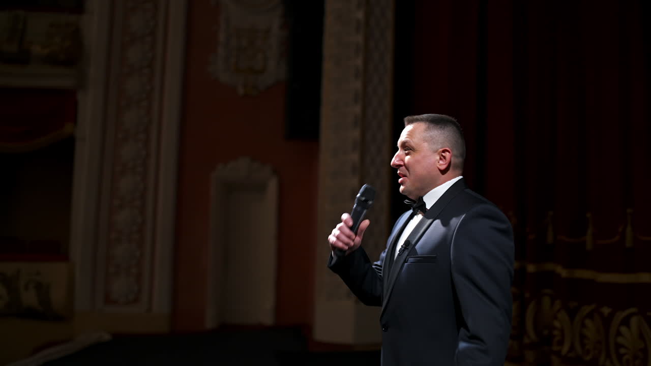 Host in elegant suit starts his speech. Speaker talking to the microphone on stage of a theater. Showman on red curtains background.
