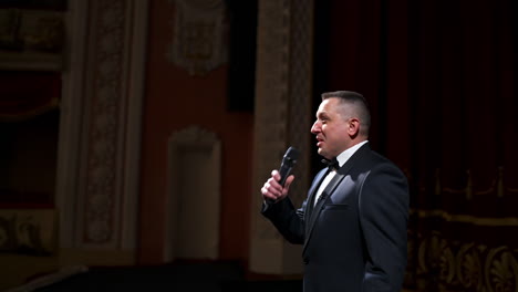 host in elegant suit starts his speech. speaker talking to the microphone on stage of a theater. showman on red curtains background.