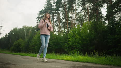 young woman in pink hoodie and jeans walking alone along tarred road through lush green forest, holding bag strap thoughtfully while surrounded by tall trees and cloudy sky on a calm quiet day