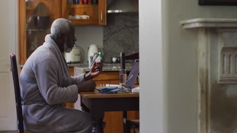hombre mayor afroamericano sosteniendo una medicación vacía mientras hace una videollamada en una computadora portátil en casa