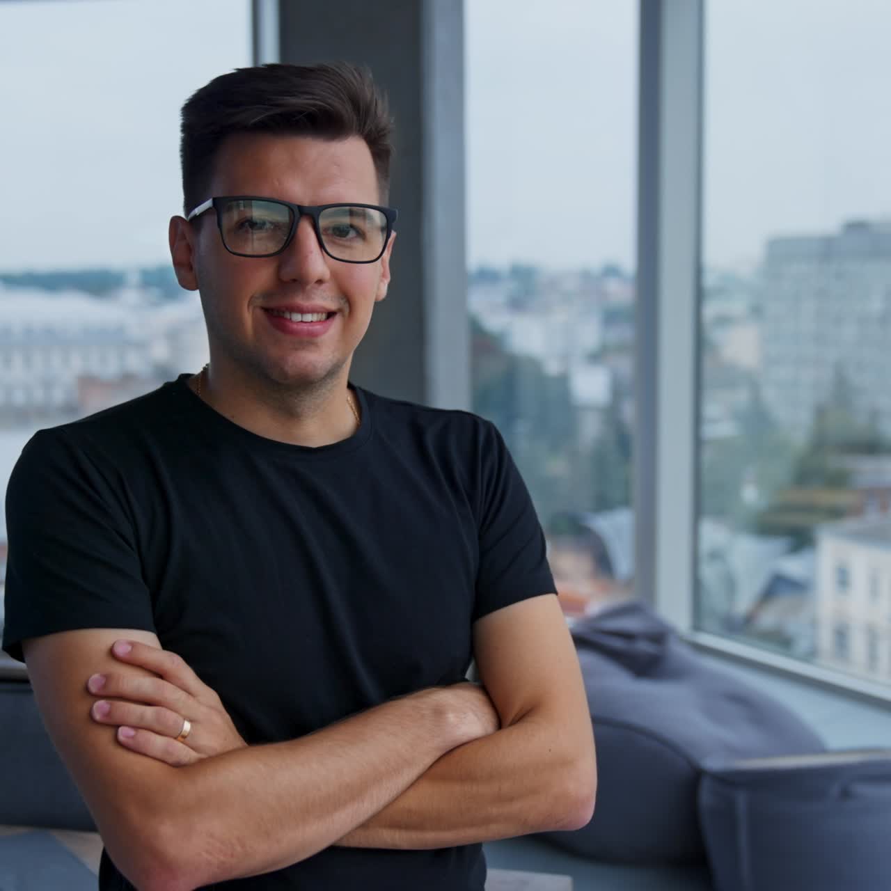 Young dark-haired man leaned on the desk in big light office. Male employee puts on glasses smiling to the camera. City view in the windows at backdrop in blur