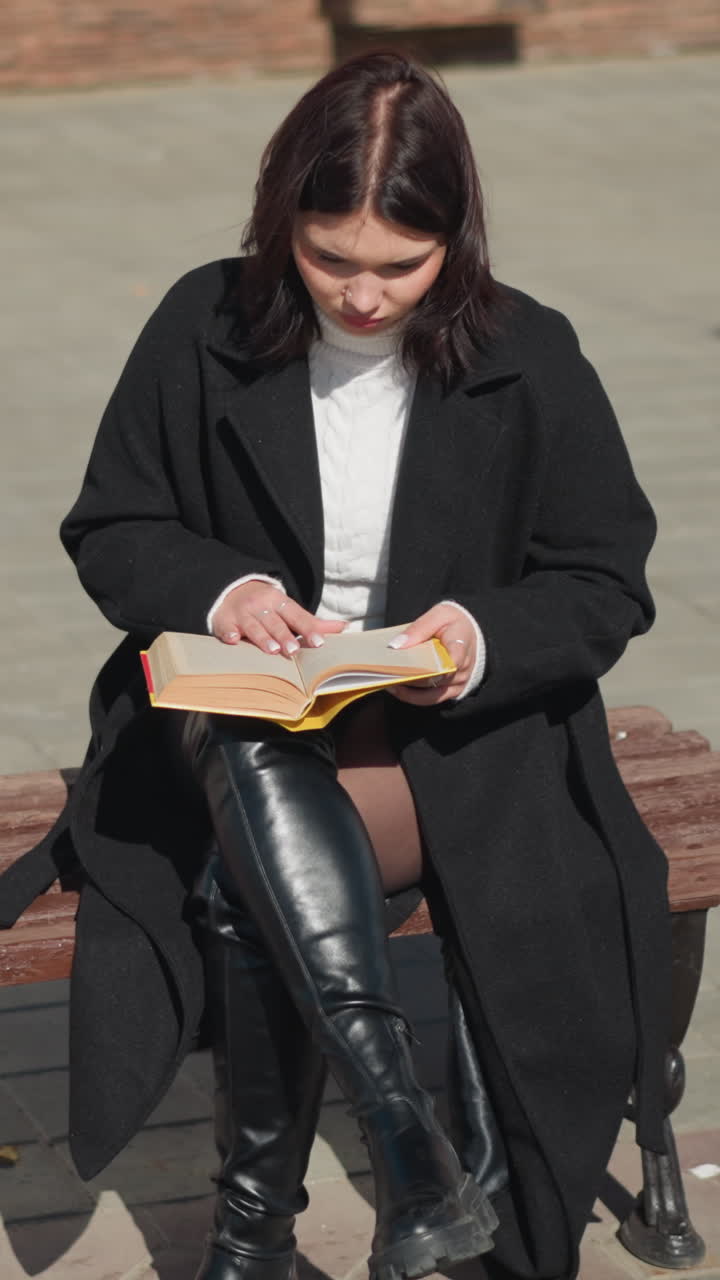 primer plano de una mujer joven leyendo un libro en un día soleado, sentada en un banco en un entorno urbano, su atención está en el libro, con su cabello y el fondo suavemente borroso