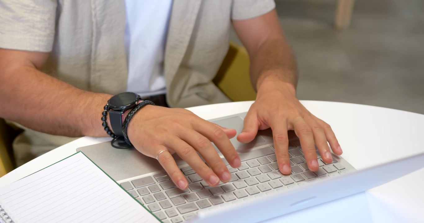 Hombre adulto comenzando a trabajar, escribiendo en una laptop plateada en una mesa redonda con un cuaderno y un reloj inteligente.
