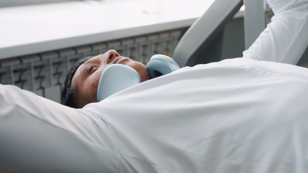 close up of man in white jacket with headphones around neck lying back on bench press machine preparing to lift barbell, focus on facial expression and upper body alignment in bright gym setting