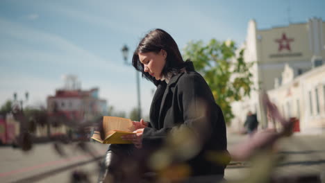 young woman in black coat sitting outdoors on a bench reading a book with focus, urban buildings and trees in the sunny background, soft sunlight illuminating her thoughtful expression