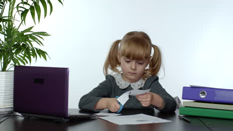 niña escolar aprende lecciones en casa sentada en la mesa cortando con tijeras formas de papel