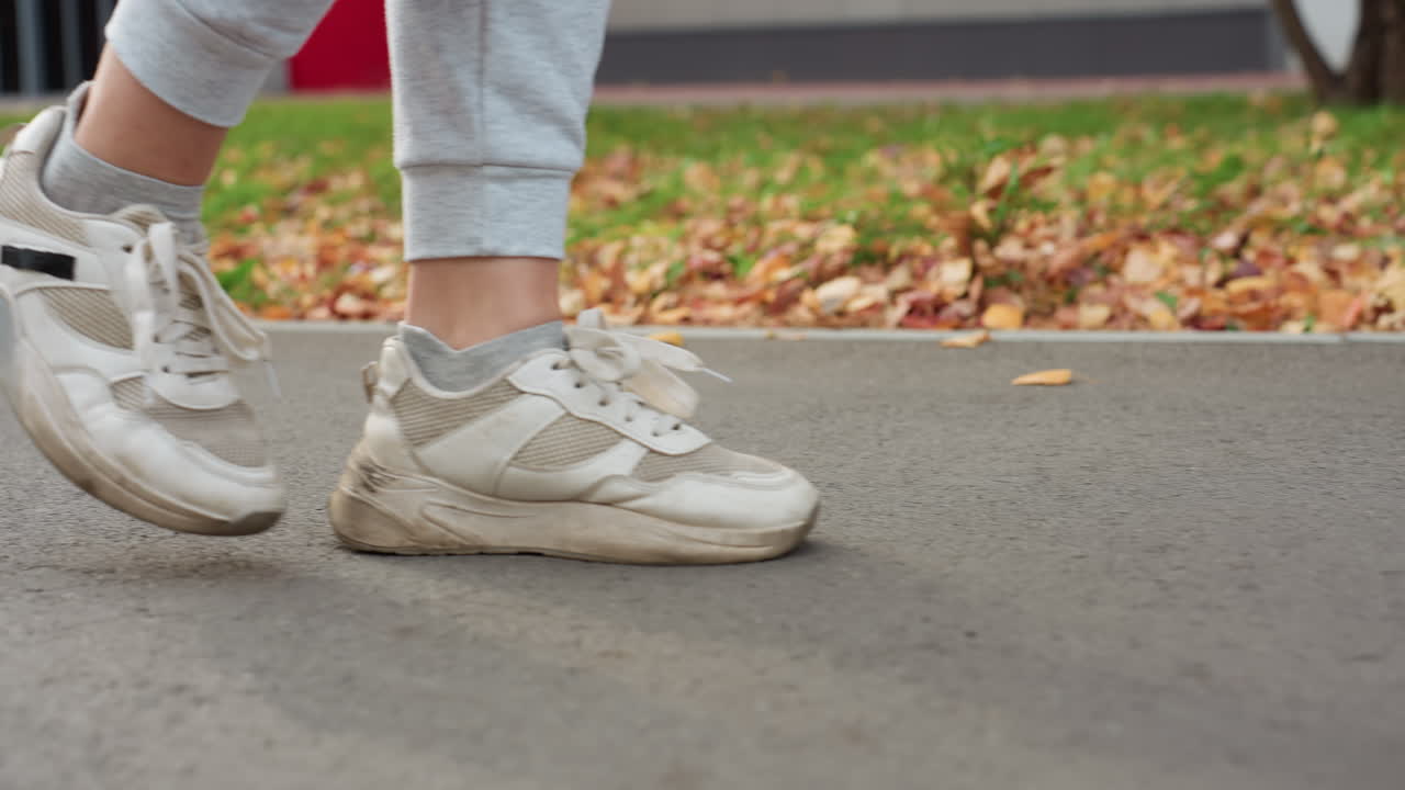Side leg view of sports person walking briskly along asphalt path with dry leaves scattered on ground swaying gently in wind while partial view of building and greenery appear in soft background blur