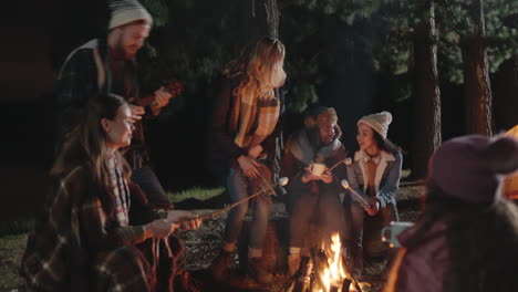 Amigos-Felices-Acampando-Sentados-Junto-A-Una-Fogata-En-El-Bosque-Por-La-Noche-Charlando-Bebiendo-Chocolate-Caliente-Asando-Malvaviscos-Compartiendo-Calidez-Disfrutando-De-Una-Aventura-Al-Aire-Libre