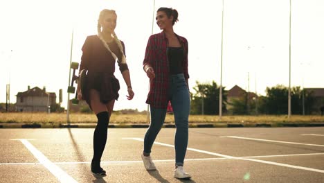 felices novias hipsters jóvenes con estilo caminando juntas en el estacionamiento. una chica con trenzas largas está sosteniendo un skateboard en