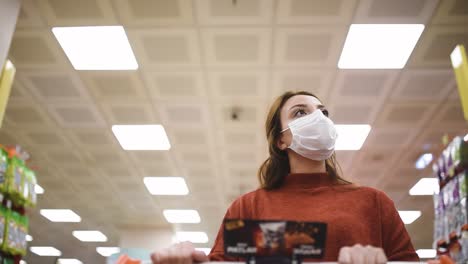 Beautiful-woman-wearing-protective-disposable-medical-mask-and-fashionable-clothes-uses-shopping-cart-while-shopping-in-supermarket