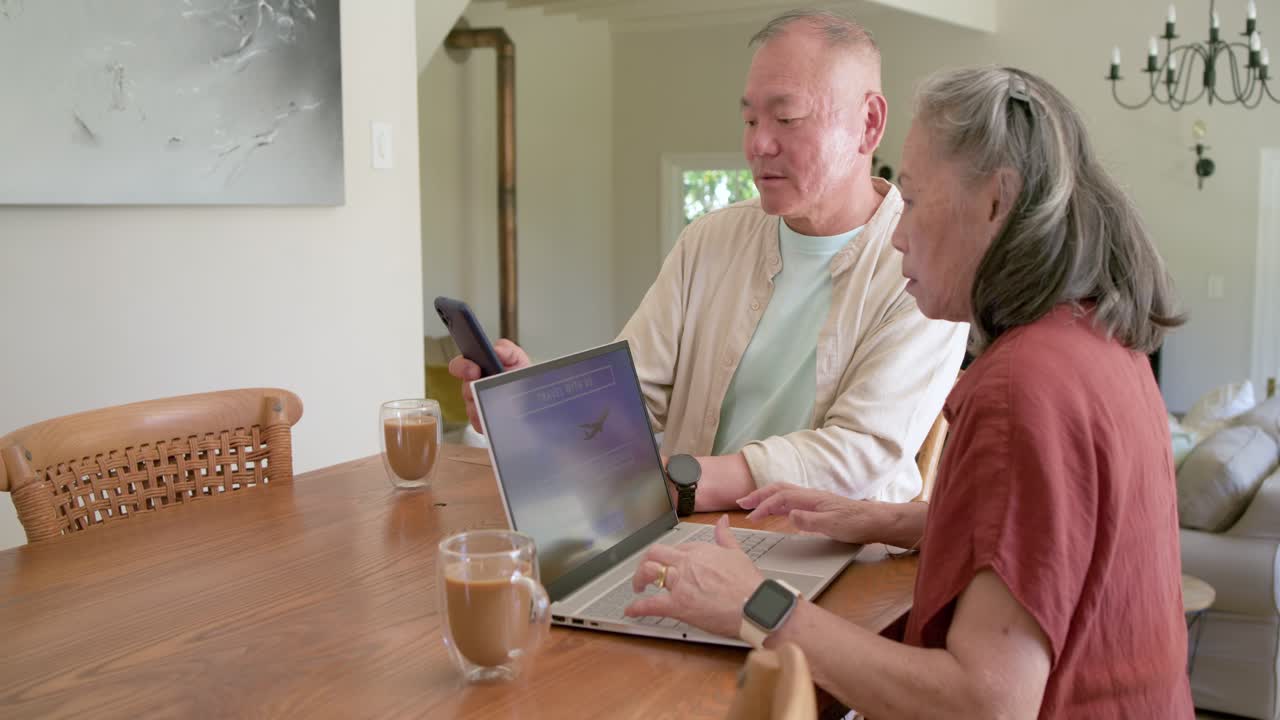 Senior Asian couple showing smartphone, typing laptop at home, finalizing flight booking with card