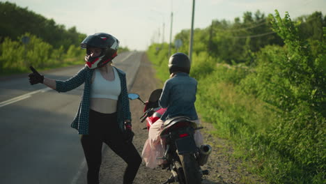 two women wearing helmets are on a roadside near greenery, one is sitting on a red sports bike while the other stands next to the bike, giving a thumbs-up, a car pass by in the background