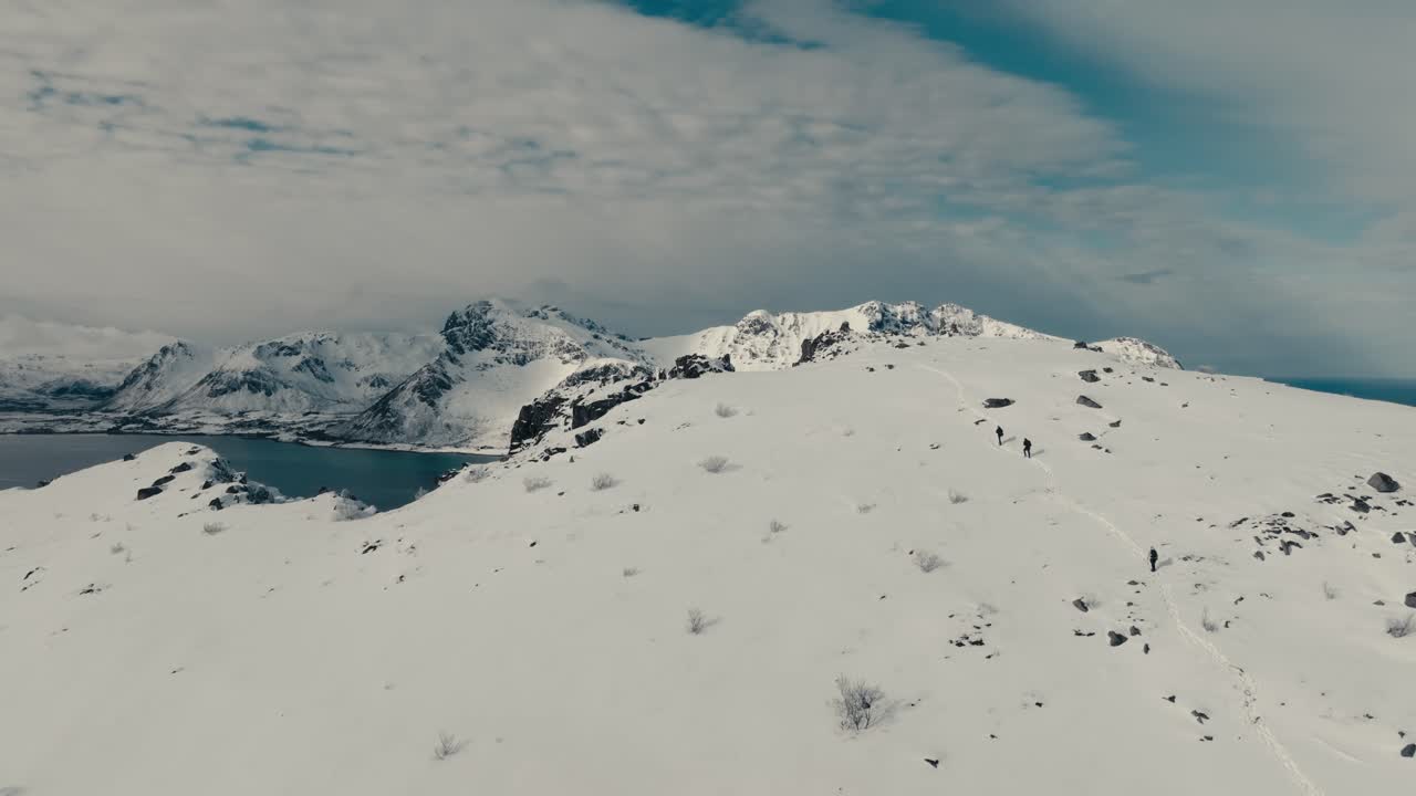 Gimsoy Beach, Lofoten Islands, Norway - Three People Hike Along a Snowy Mountain Trail Overlooking a Stunning Fjord and the Rugged Peaks - Pan Up Shot