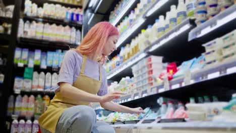 Side-view-of-a-confident-girl-with-pink-hair-working-in-a-supermarket-and-placing-goods-on-the-dairy-products-counter-in-a-supermarket