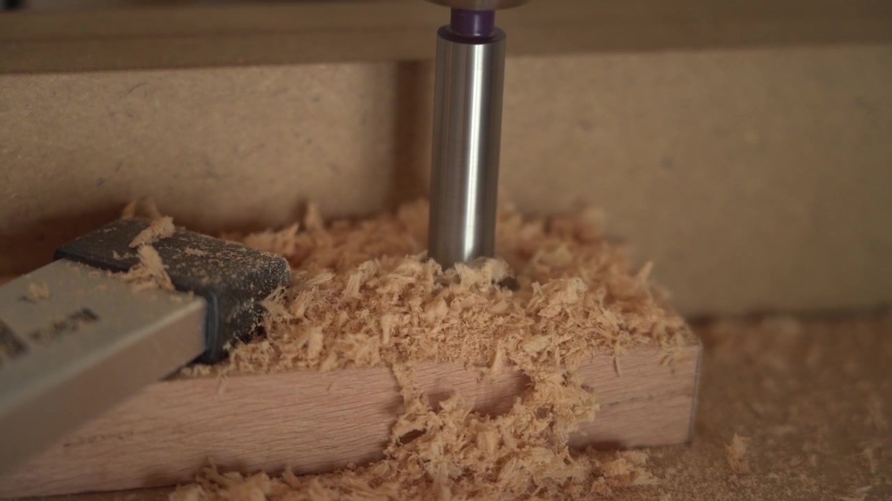 Closeup of router bit cutting wooden board, removing timber to form clean recess while wood chips collect during woodworking process on workbench