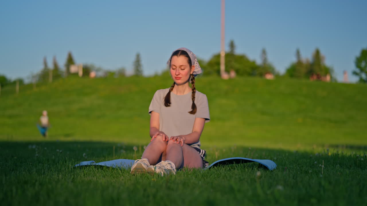 A young woman sits on her yoga mat with a stunning skin tone highlighted by the golden evening sun. A green hill rises behind her. She feels a complete a well-being. Camera slides to the right.